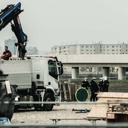 Ein Bagger reißt die selbstgebauten Unterstände des Protestcamps in der Hausfeldstraße nieder. Die Polizei sieht aus der Distanz zu.