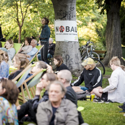 Publikum bei Wien am Fluss, auf einem Baum hängt ein "Nobau" Plakat